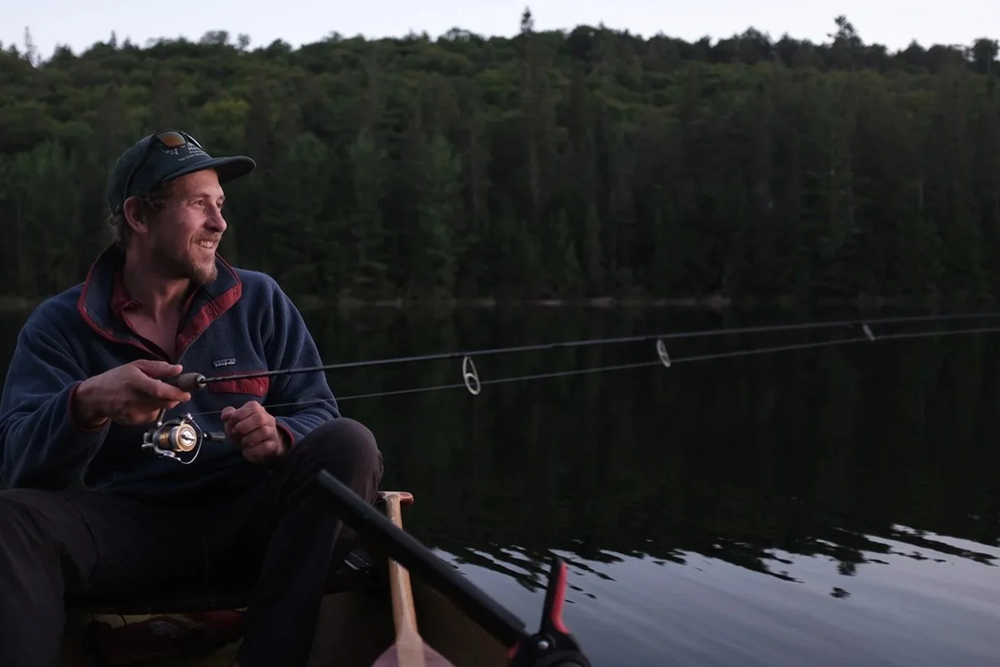Man fishing on a boat during sunset on a calm lake surrounded by dense forest.