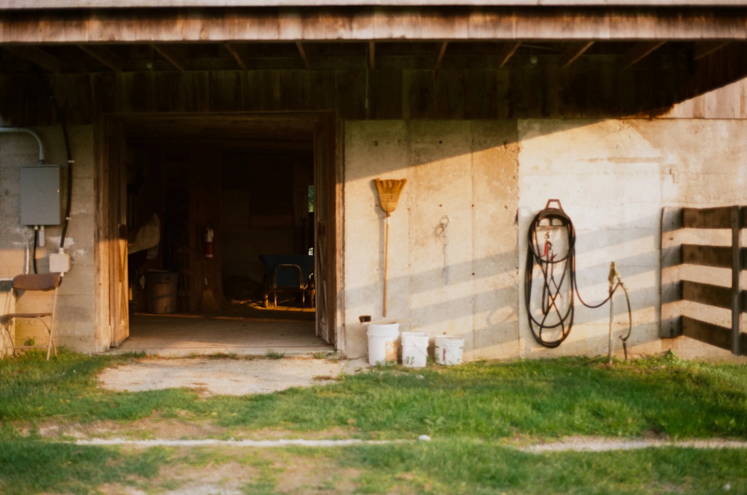 Exterior view of a garage with open door, showing some outdoor tools hanging on the wall, a broom, a horse saddle, and buckets outside on grass.