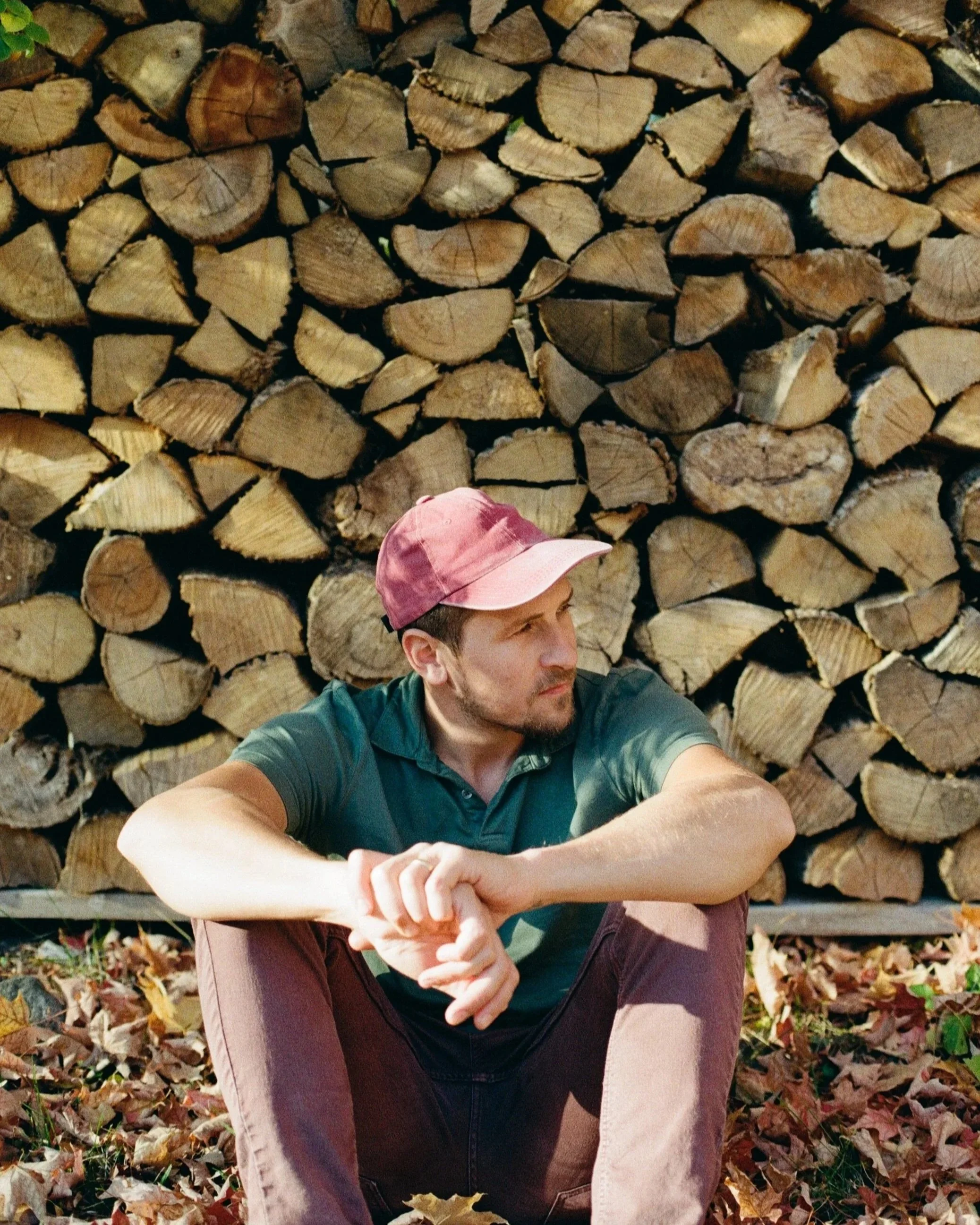 A man in a green shirt and maroon pants sitting on the ground in front of a large stack of cut wood logs, wearing a pink cap.