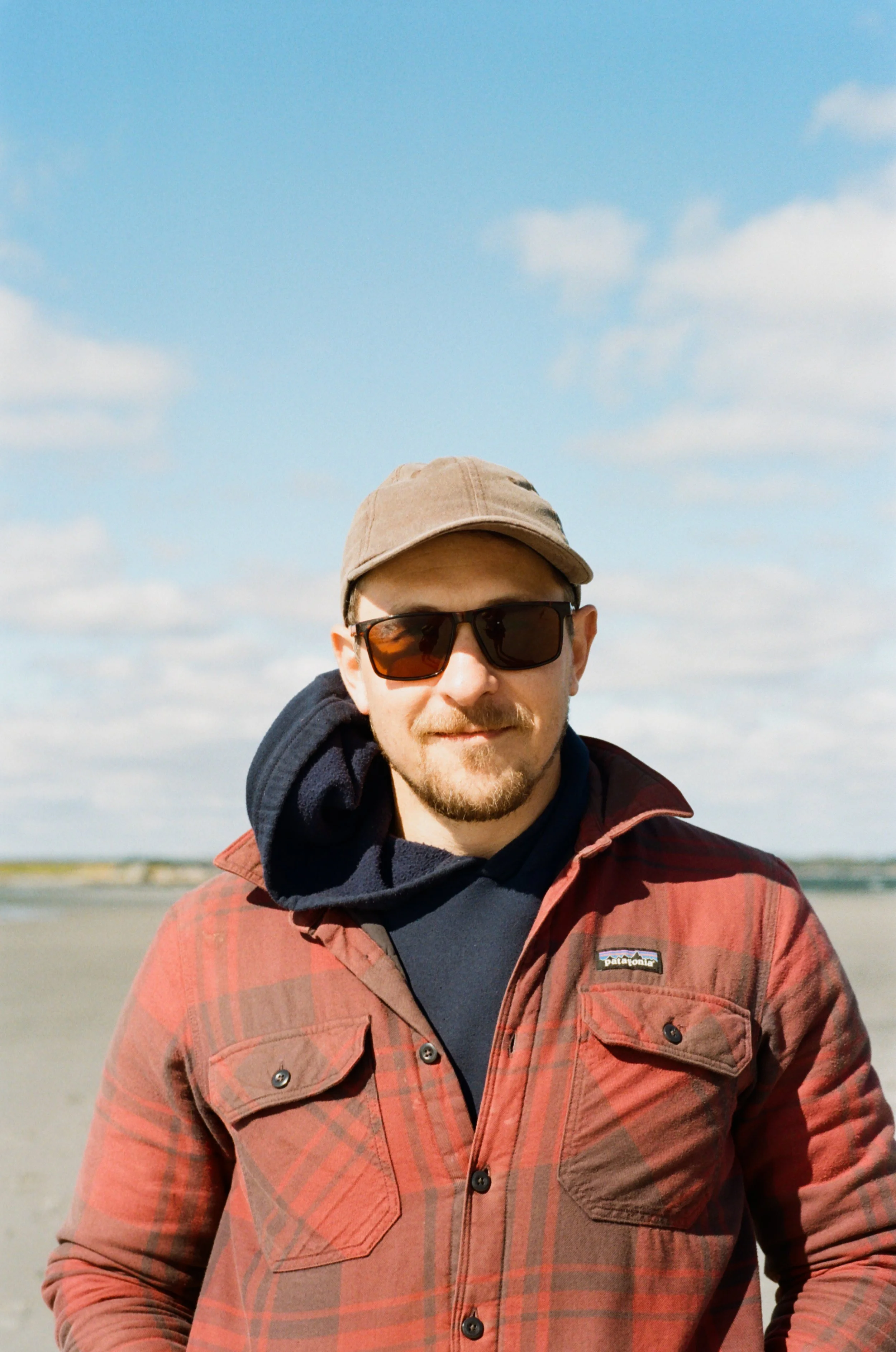 A man wearing sunglasses, a beige cap, and a red plaid jacket standing outdoors against a sky with some clouds.