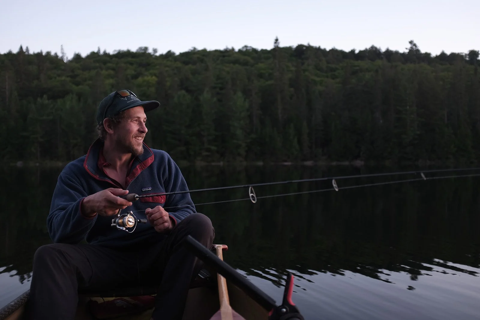 A man fishing in a canoe on a lake surrounded by trees during dusk, smiling and wearing a cap and fleece jacket.