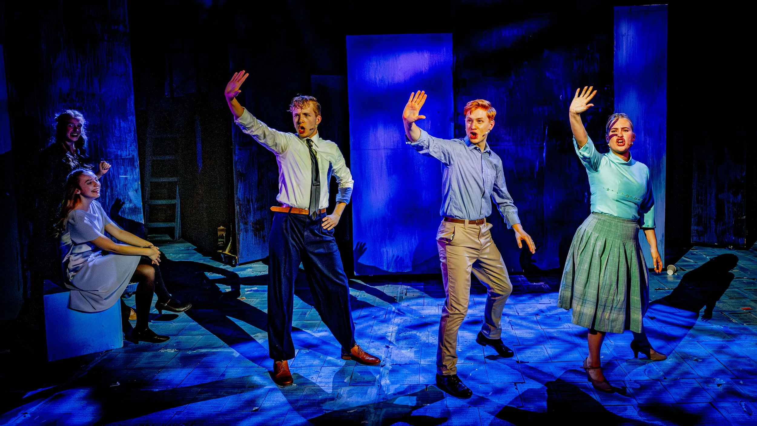 Group of five young actors performing on stage with blue lighting, three standing and two seated, in a dramatic scene with expressive gestures and colorful costumes.