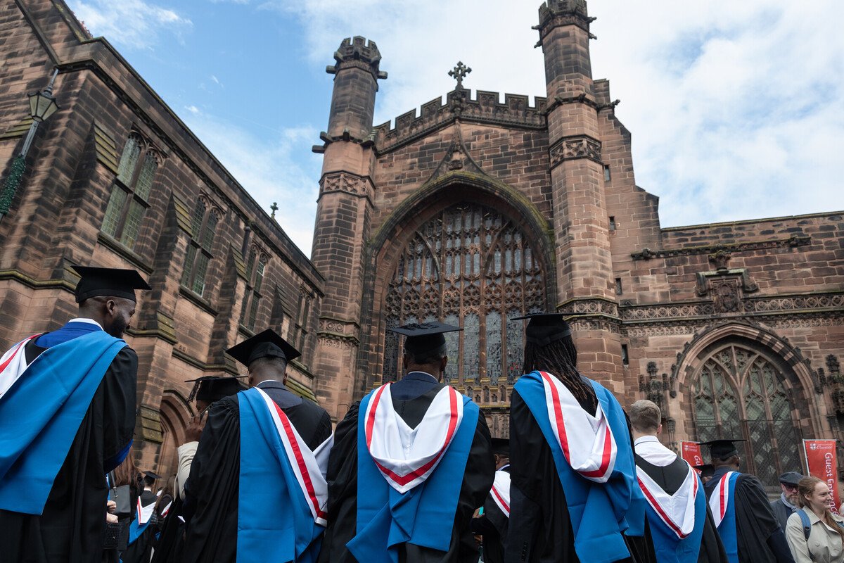 Graduates in caps and gowns standing outside a historic stone building with large stained glass windows, likely a church or academic institution.