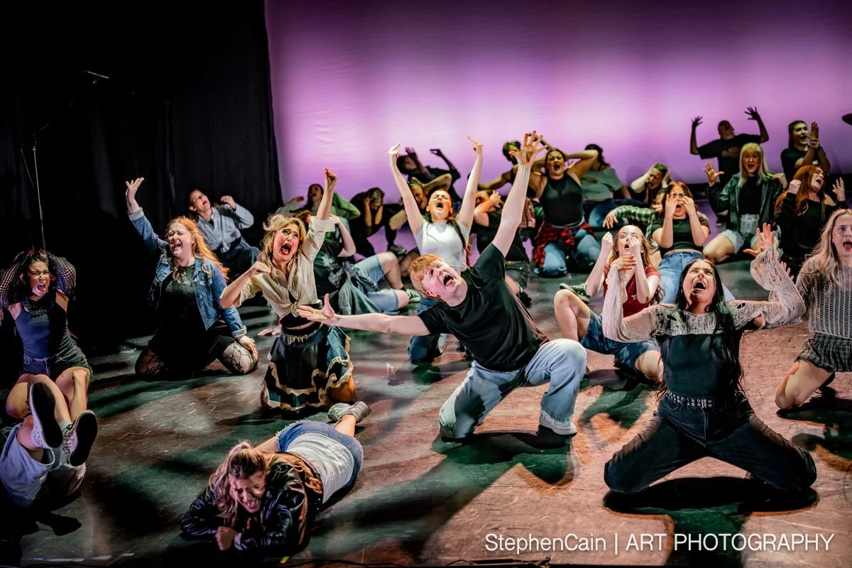 A large group of young people on stage performing a theatrical dance or musical with expressive gestures and colorful costumes, backlit with purple lighting.