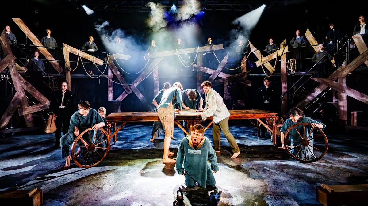 Scene from a theatrical play with actors on stage. The stage has a large wooden table, and actors are engaged in a dramatic scene, with some leaning over the table and others pushing a wagon. The background has wooden structures and rigging, with additional actors standing on elevated platforms. The lighting is intense, highlighting the performers and creating a dramatic atmosphere.