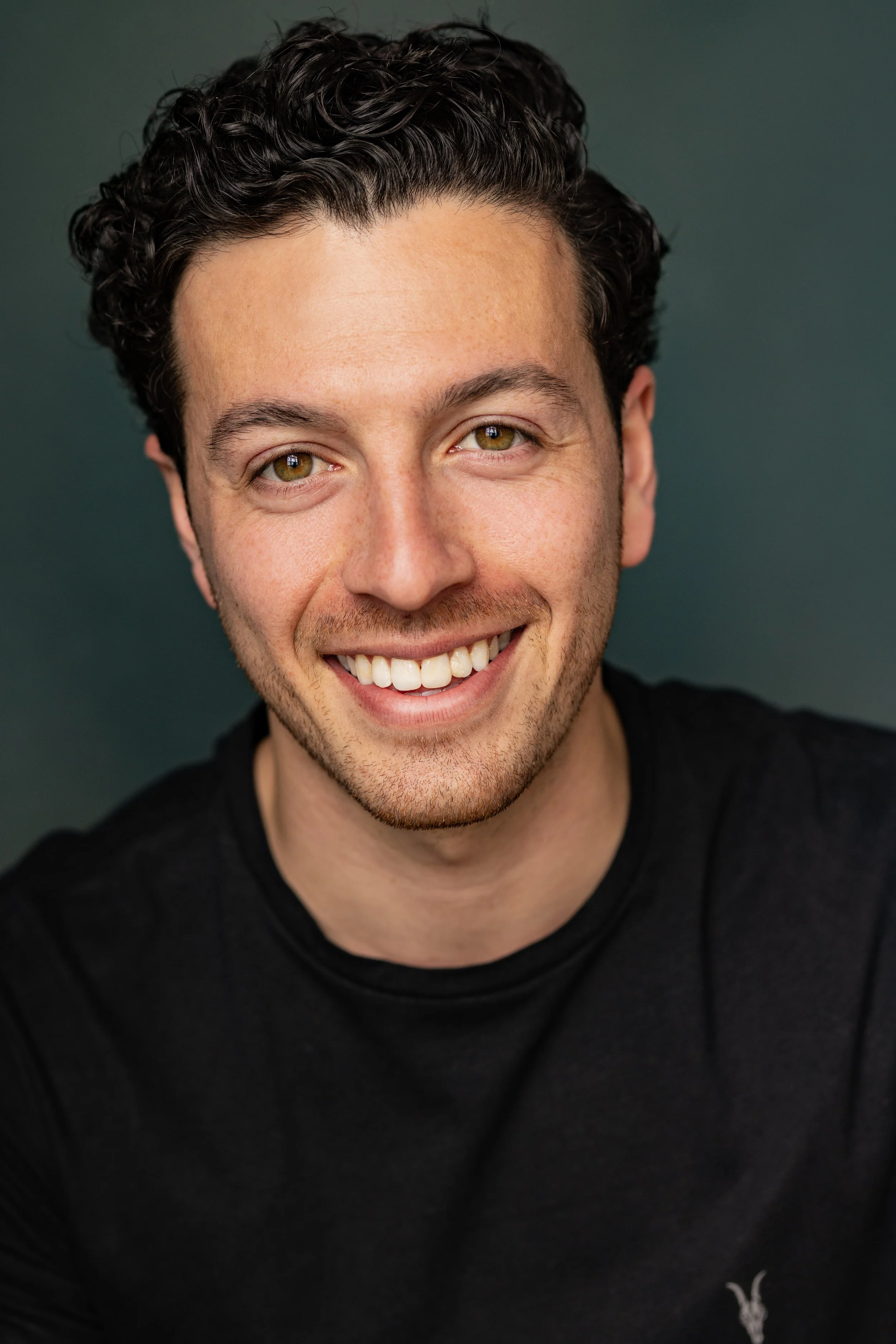 Close-up of a smiling man with curly dark hair, hazel eyes, wearing a black shirt, against a dark teal background.