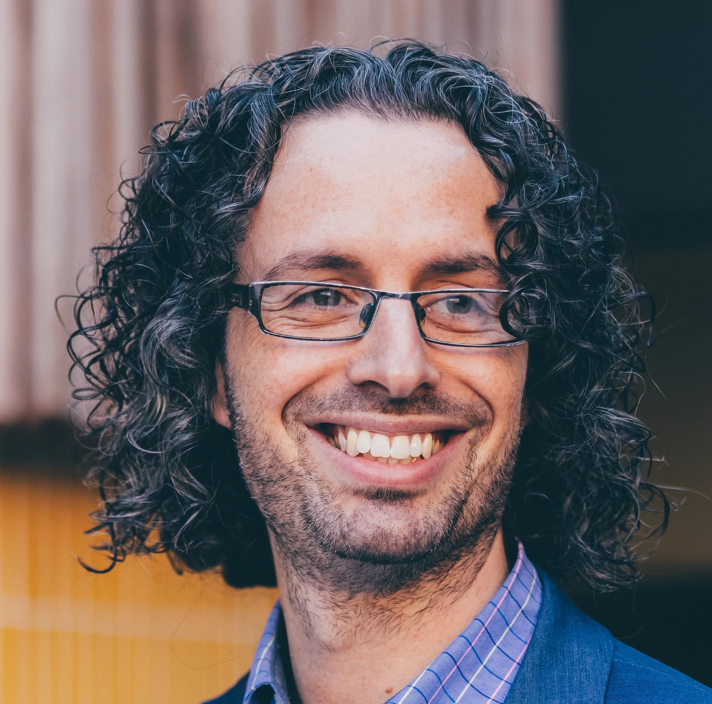 A close-up of a person with curly dark hair, glasses, and a beard smiling, wearing a blue checkered shirt.