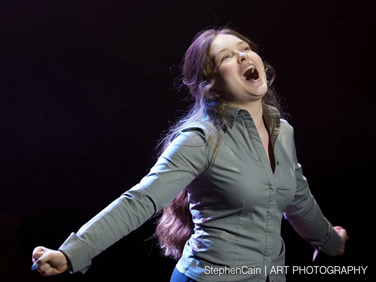 A woman with long, wavy hair singing passionately with her arms outstretched against a dark background.