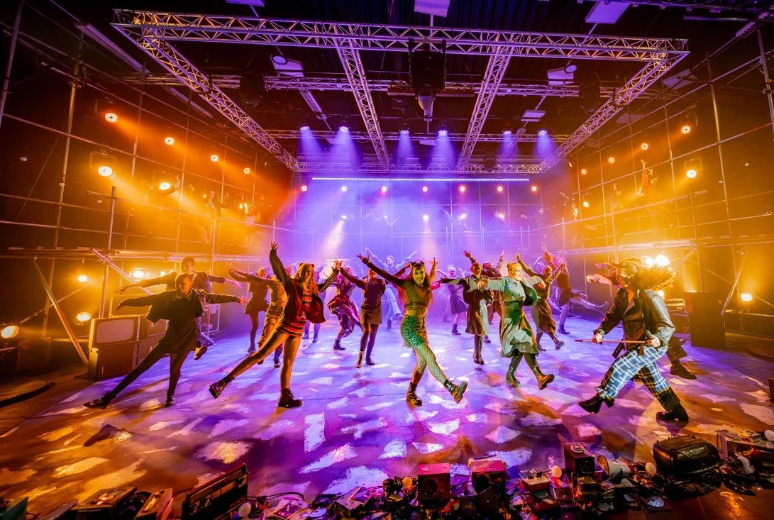 Dance performers on stage with colorful lighting in a large studio or theater with a metal scaffolding ceiling.