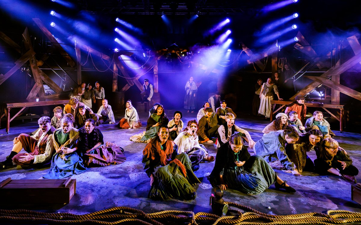 A theatrical stage with a large cast of performers in period costumes, performing a dramatic scene under colorful stage lighting with a rustic set design.