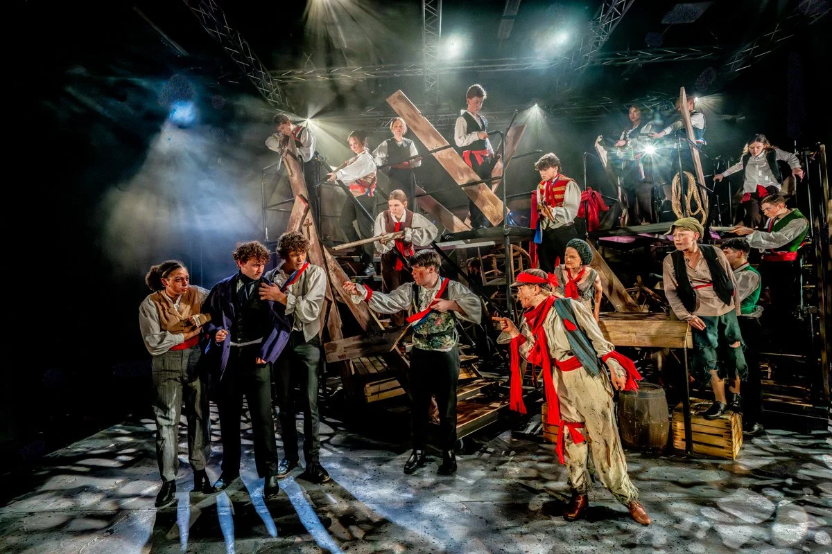 A theatrical stage filled with actors dressed in military-style costumes, with some holding props like guns, surrounded by wooden structures and scaffolding, under dramatic stage lighting.