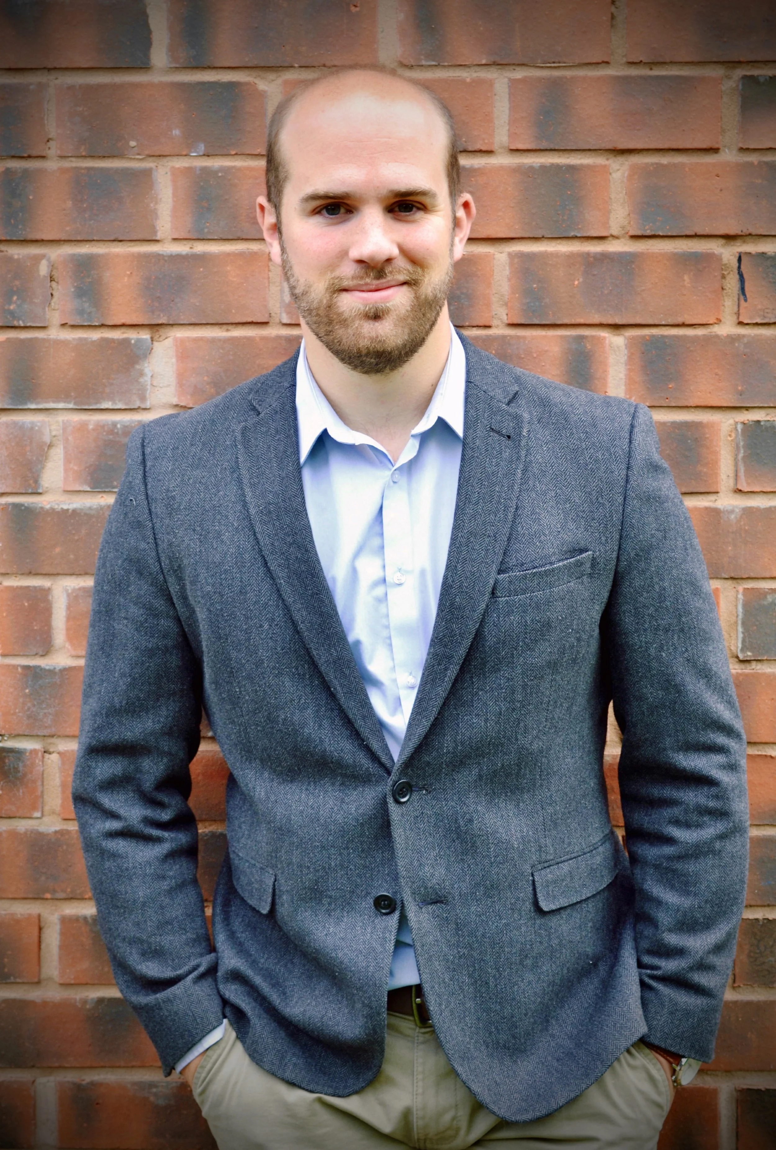 A man with a beard and short hair, dressed in a gray blazer and white shirt, standing in front of a brick wall, with his hands in his pockets, smiling slightly.