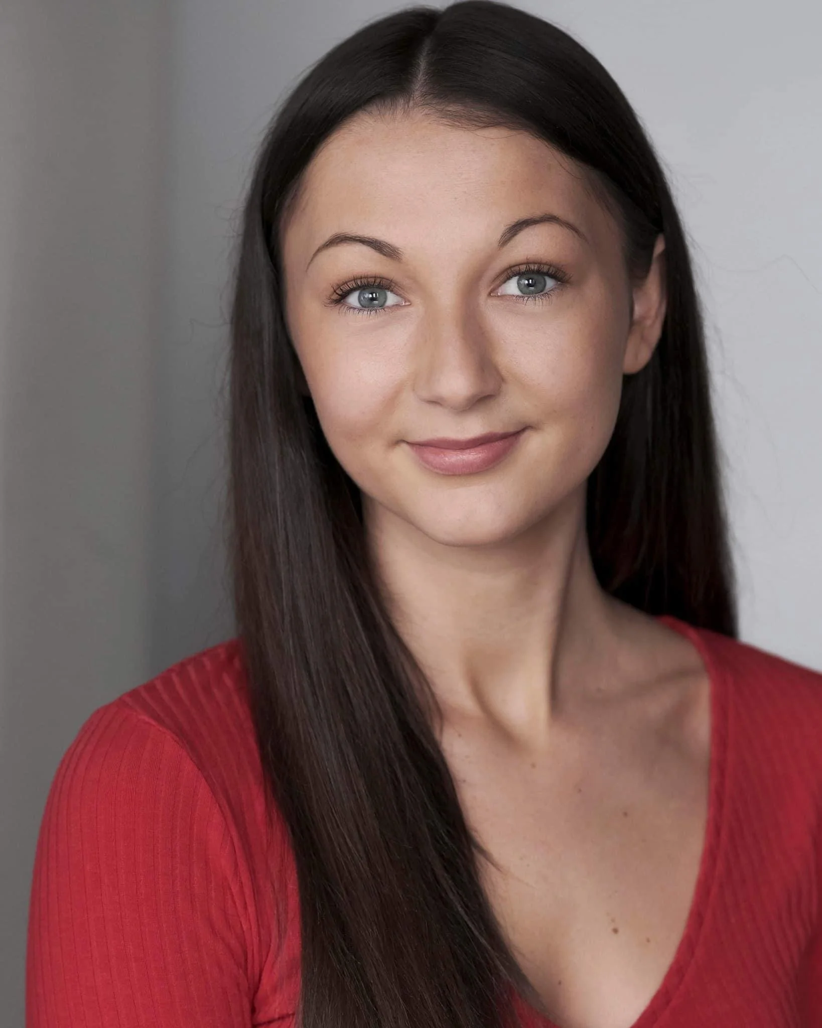 Close-up portrait of a young woman with long dark hair, blue eyes, wearing a red top, smiling softly, with a neutral background.