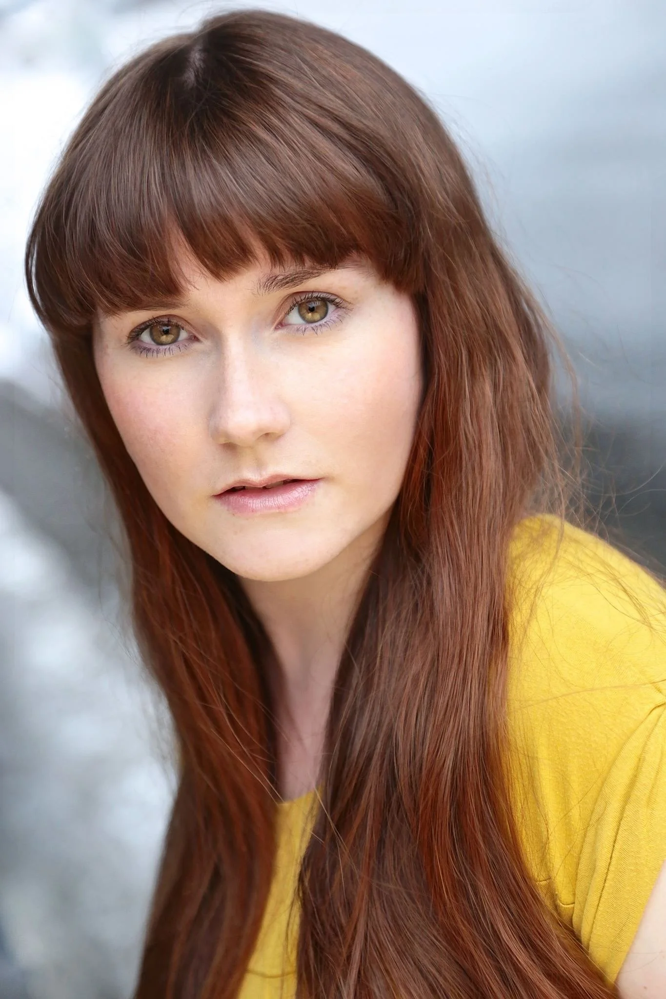 Close-up portrait of a young woman with hazel eyes, straight auburn hair with bangs, wearing a yellow top, looking at the camera.