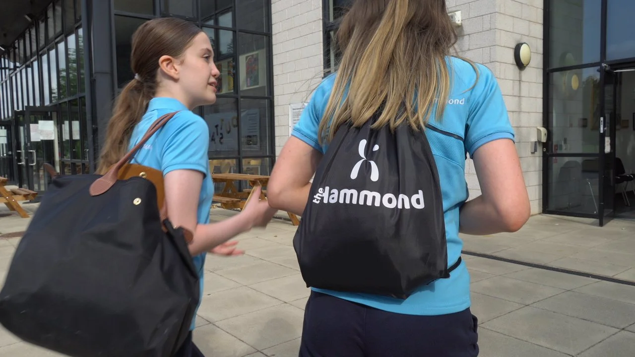 Two young girls in blue shirts walking outside a modern building with glass windows, carrying black bags with the logo 'the hamond'.