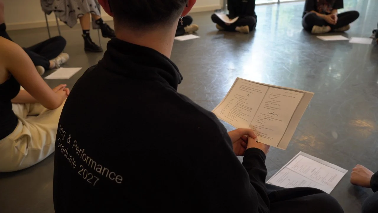 A person sitting on the floor reading a program or menu, with several other people sitting around in a circle, holding papers, in a room with a large window.