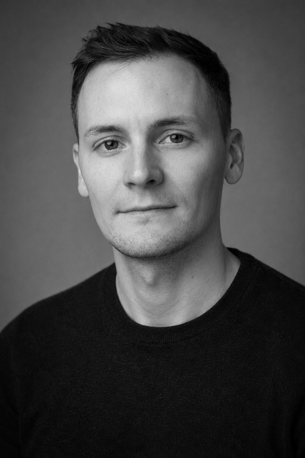 Black and white portrait of a young man with short dark hair, wearing a dark crewneck shirt, looking directly at the camera with a slight smile.