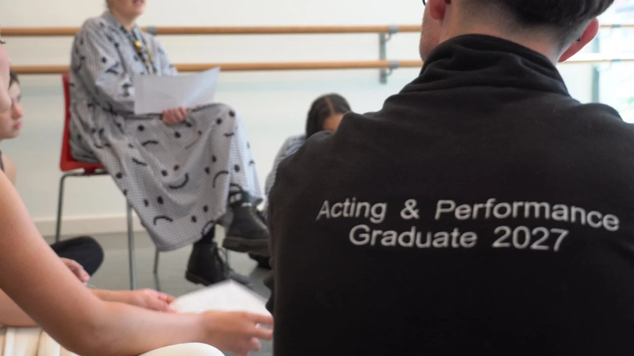 A person wearing a black hoodie with white text that reads "Acting & Performance Graduate 2027", sitting in a room with other people. An individual in black shoes and a patterned dress is sitting on a chair, holding papers, with a ballet barre on the wall behind them.
