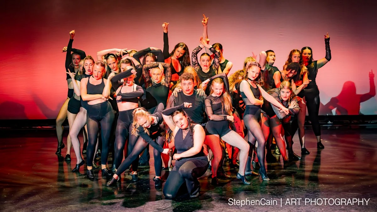 Group of young female dancers in black outfits performing on stage with dramatic poses and colorful background lighting.