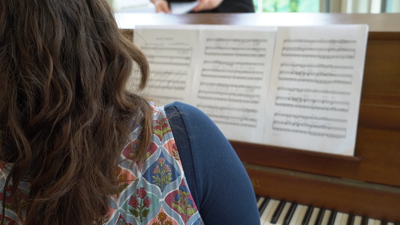 A woman with long, wavy brown hair is sitting in front of a piano, reading sheet music.