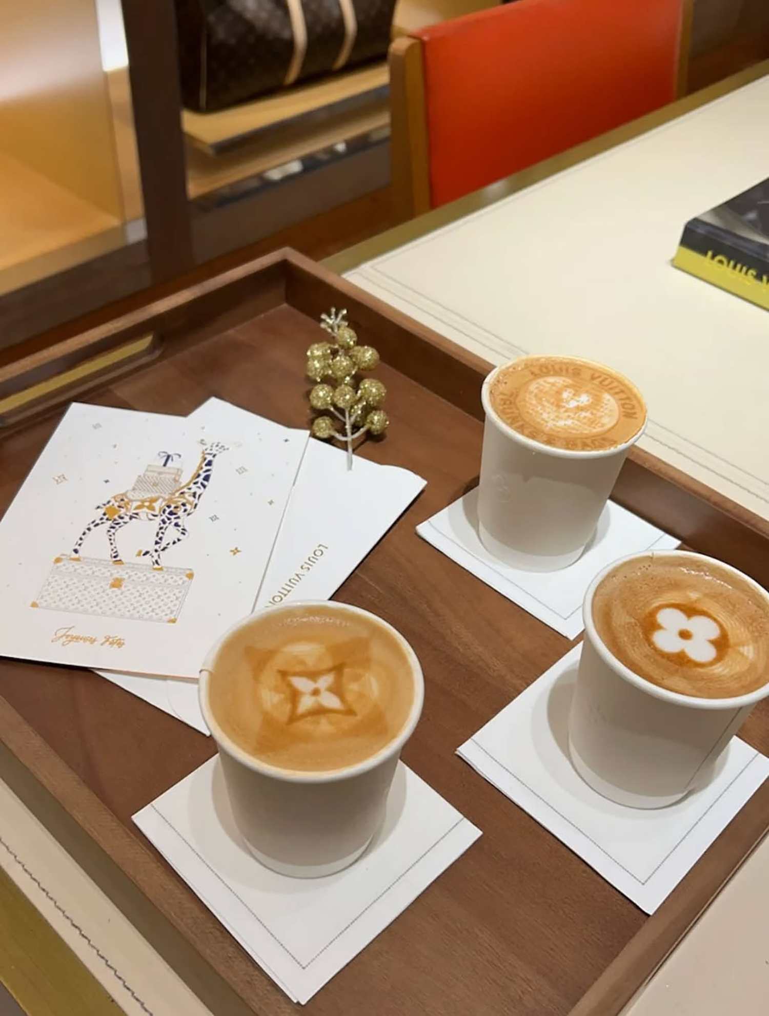Three cups of coffee with steamed milk art, on white napkins, on a wooden tray in a cafe setting, with holiday cards and a small gold decorative piece. Get award winning coffee for your retail spot from bean sprout coffee and drink catering in MN