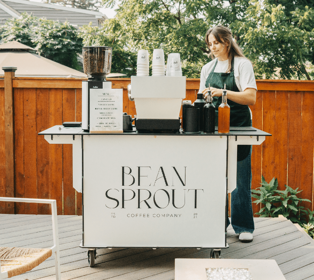 A woman standing at a coffee cart labeled 'Bean Sprout Coffee Company' in MN, preparing drinks. The cart has a menu and various bottles, with greenery and a wooden fence in the background. Get award winning coffee from bean sprout coffee catering