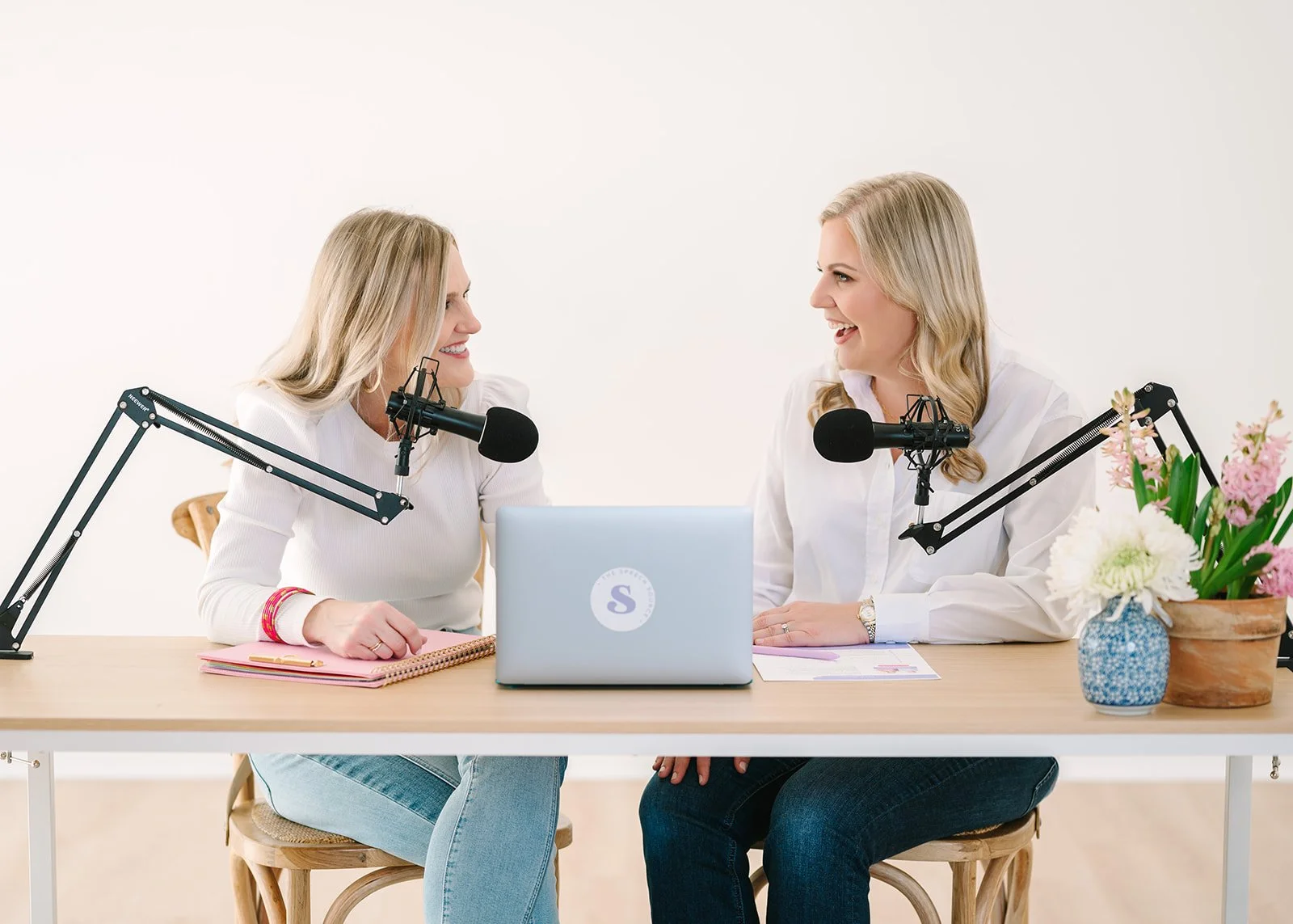 Two women sitting at a table with microphones, engaging in a podcast recording or interview. They are smiling, facing each other, with notebooks, a laptop, and flowers on the table.