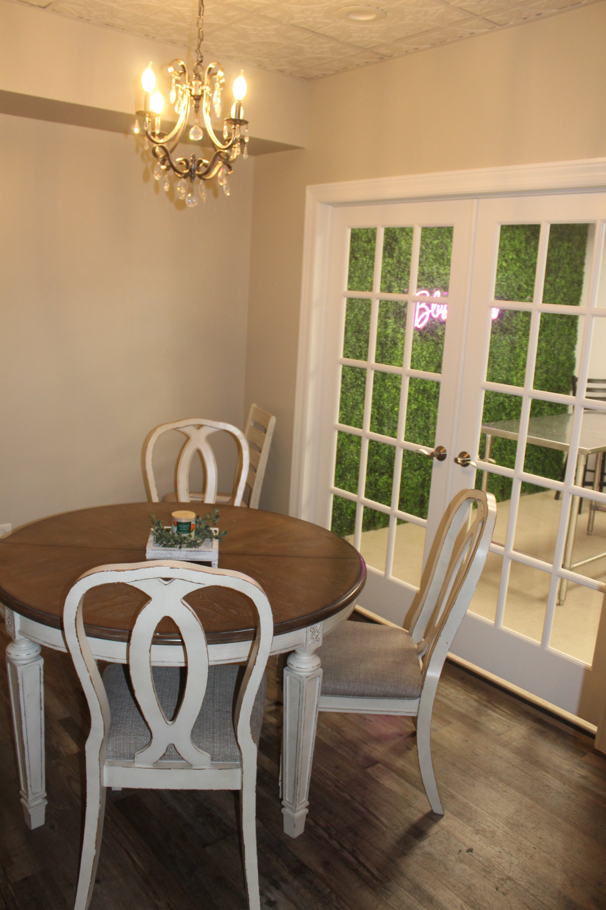 Consultation area with rustic wooden round table, white chairs, chandelier, and French doors leading to a room with green wall covering.