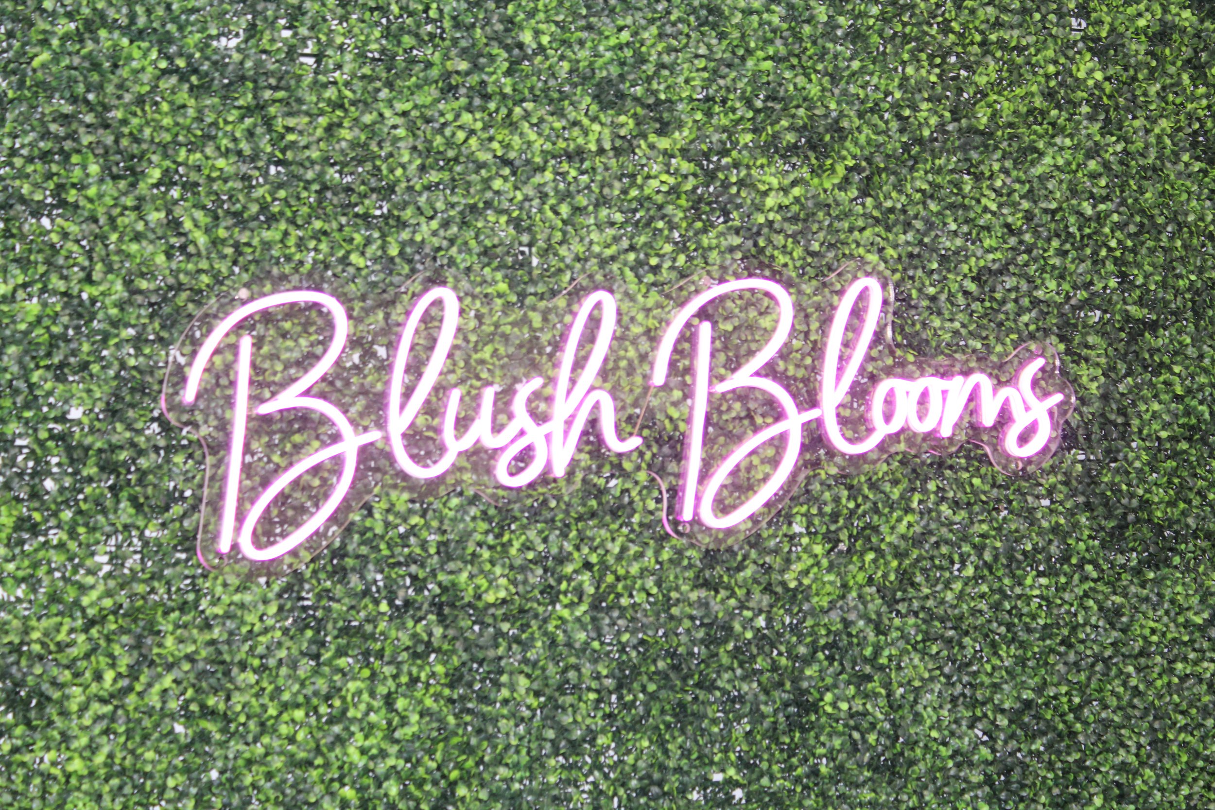 Pink neon sign "Blush Blooms" on green foliage background