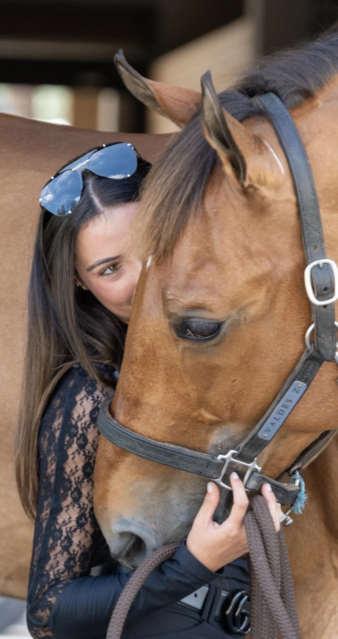 A woman with long dark hair and sunglasses on her head is hugging a brown horse. The woman's face is partly hidden behind the horse's head. The horse is wearing a bridle with a leather strap and a metal nameplate.
