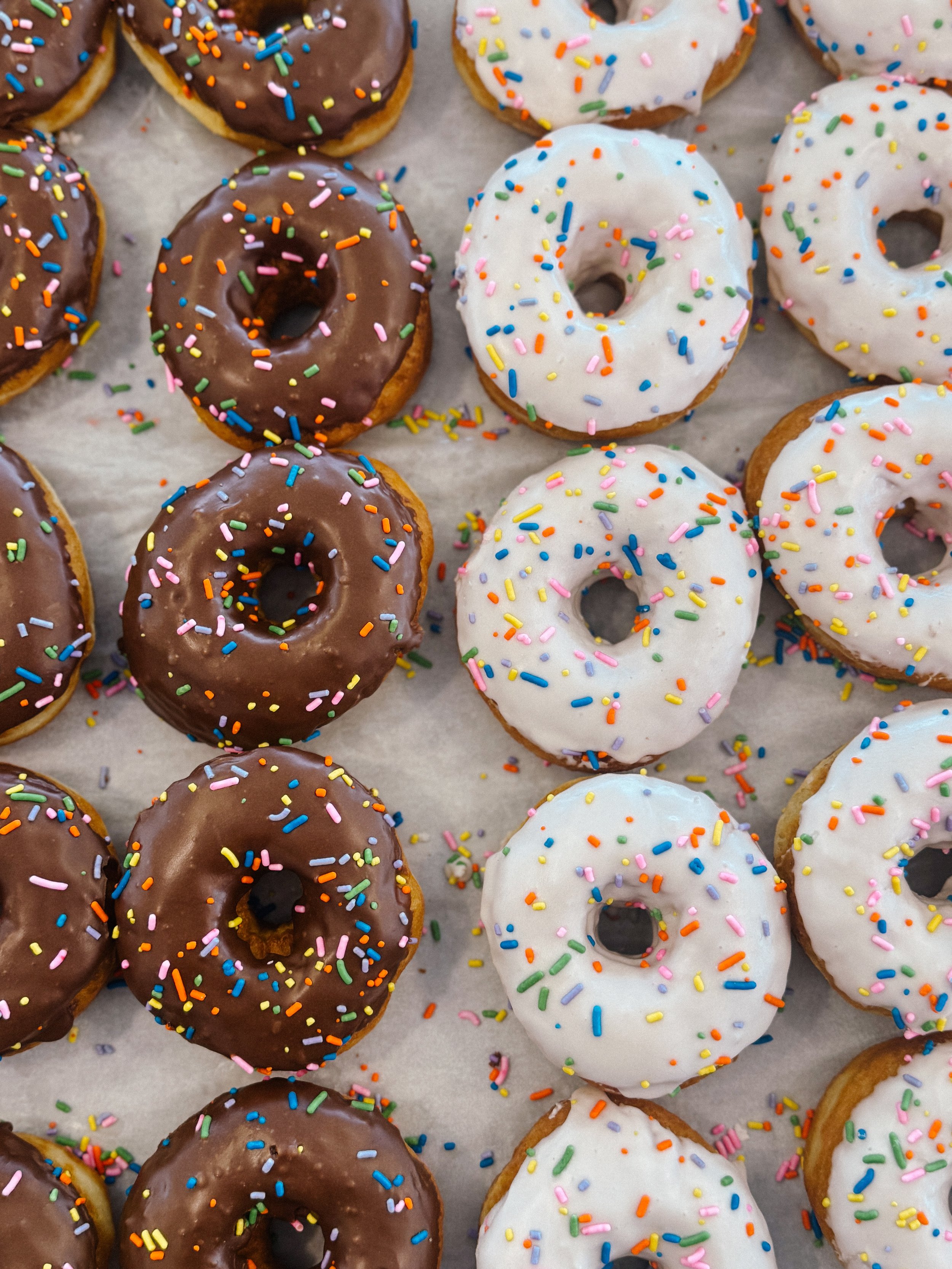 Assorted donuts with chocolate and white icing topped with colorful sprinkles, arranged on parchment paper.