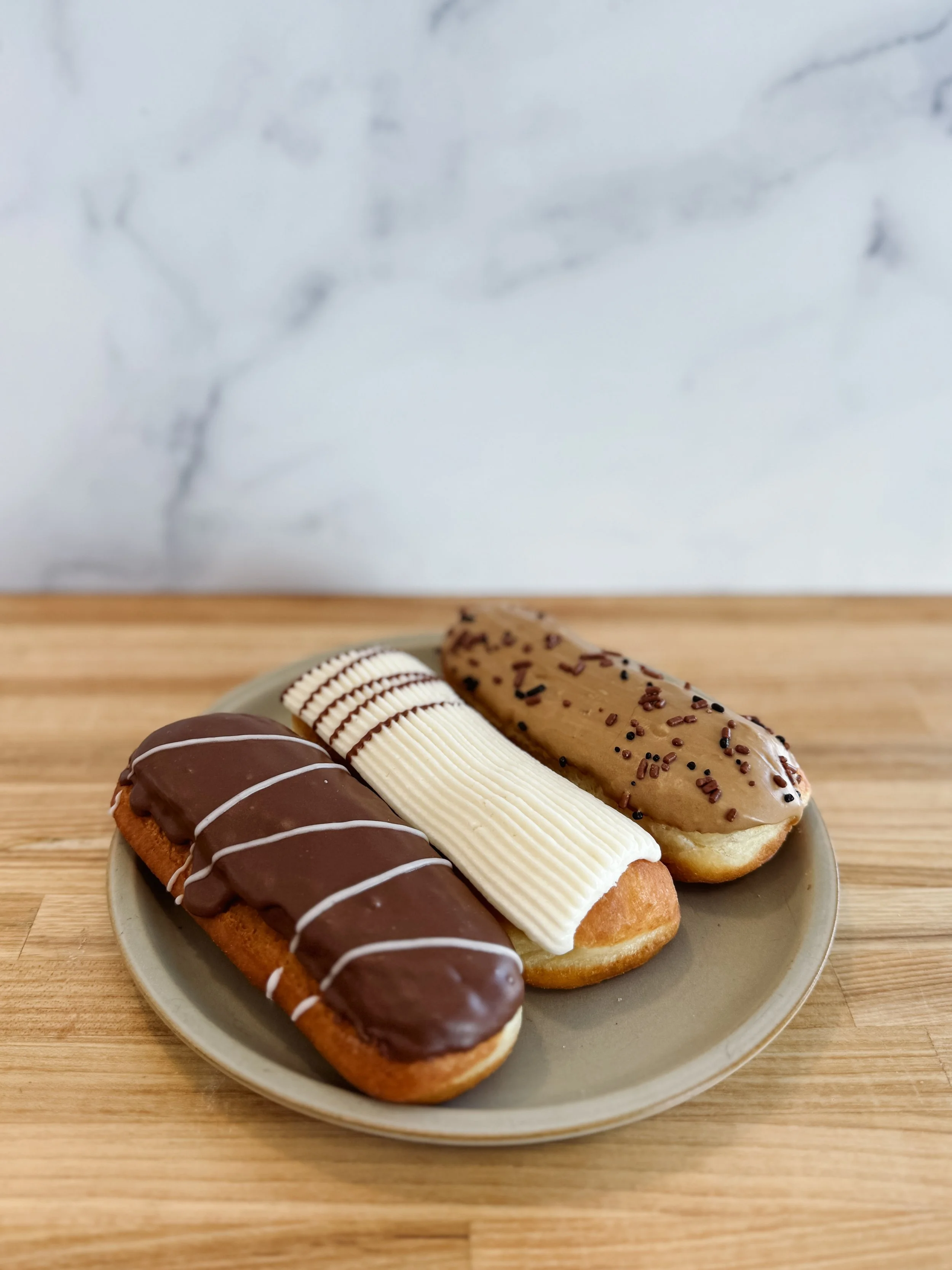 Three assorted eclairs on a small gray plate placed on a wooden surface with a white marble background. The eclairs are decorated with chocolate, white, and caramel toppings.