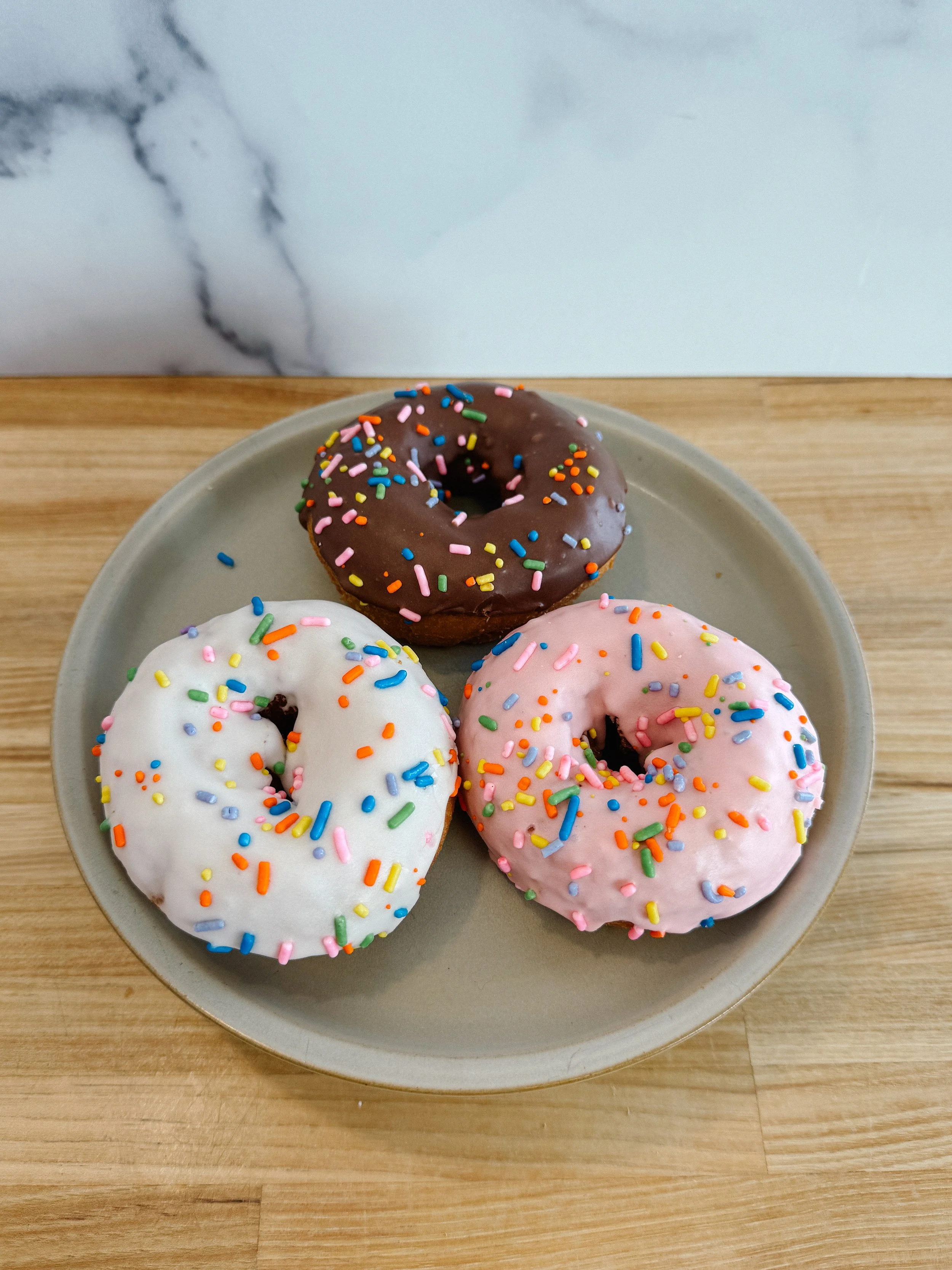 Three donuts with colorful sprinkles on a beige plate placed on a wooden surface.