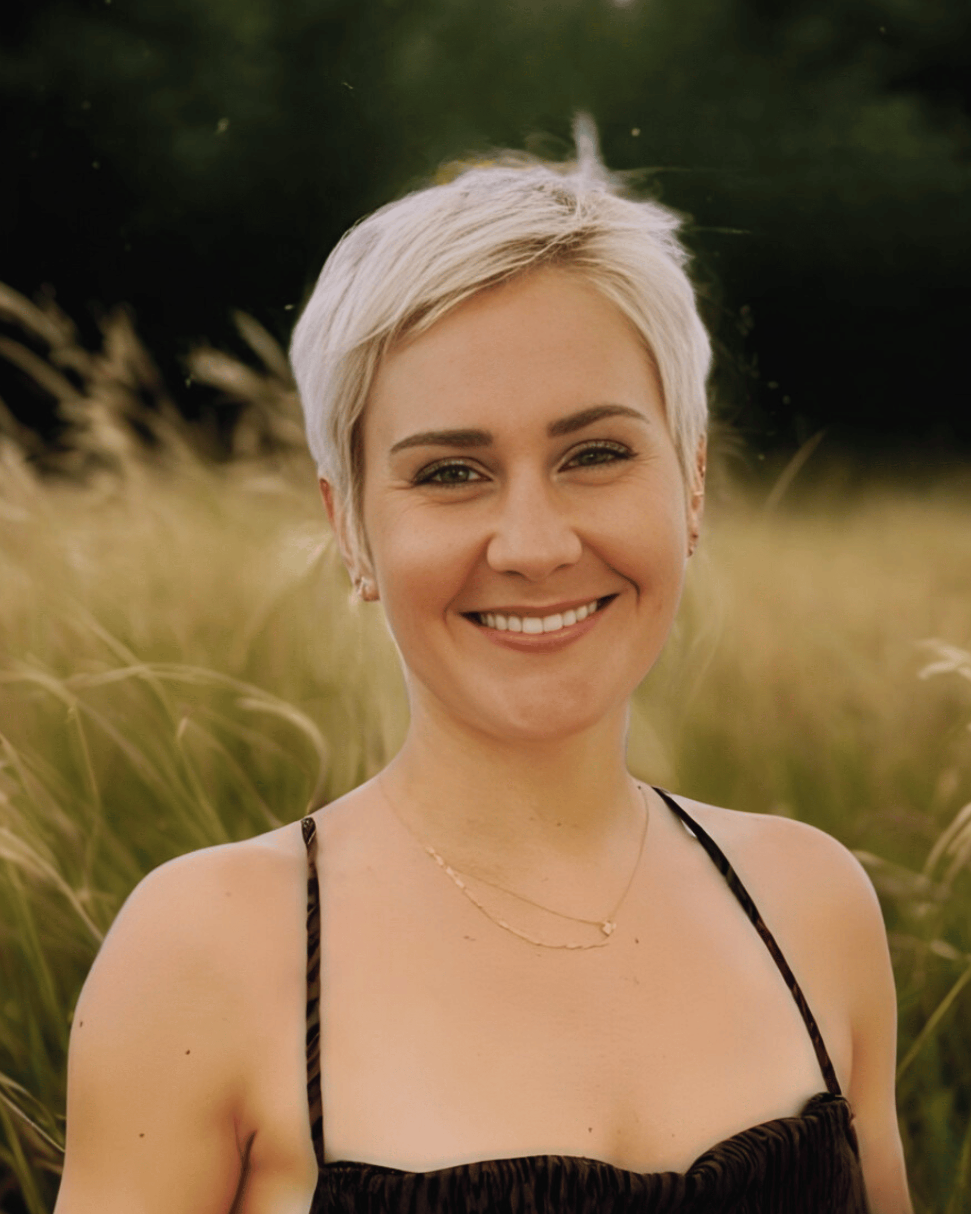 A woman with short, platinum blonde hair smiling outdoors in a field of tall grasses.