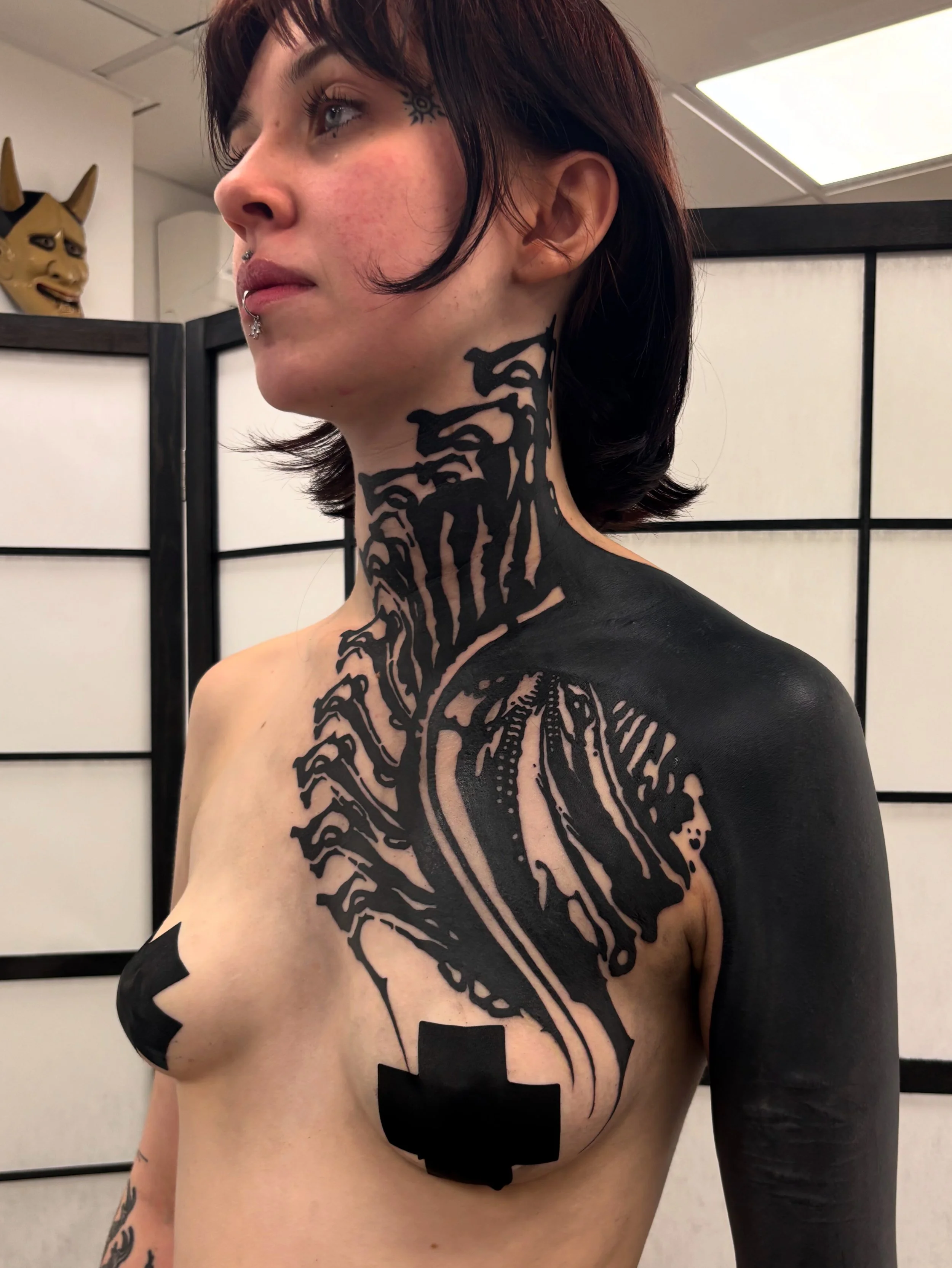 A woman with short dark hair and tattoos on her neck, shoulder, and chest, some of which are covered in black paint, stands in front of a folding screen.