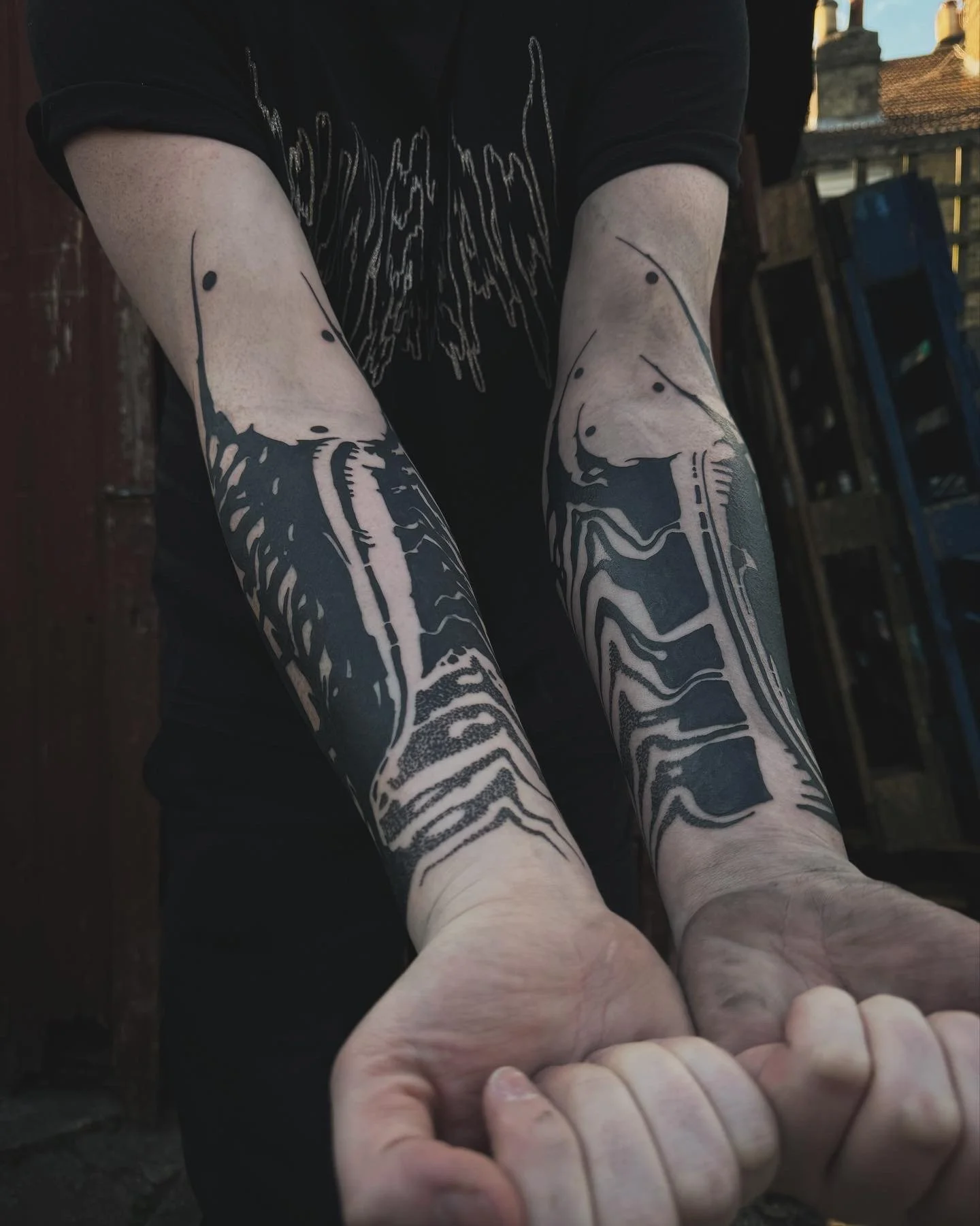 Person with black ink tattoos on both forearms, holding hands with fingers intertwined, wearing a black t-shirt with a white graphic design, in an urban outdoor setting.