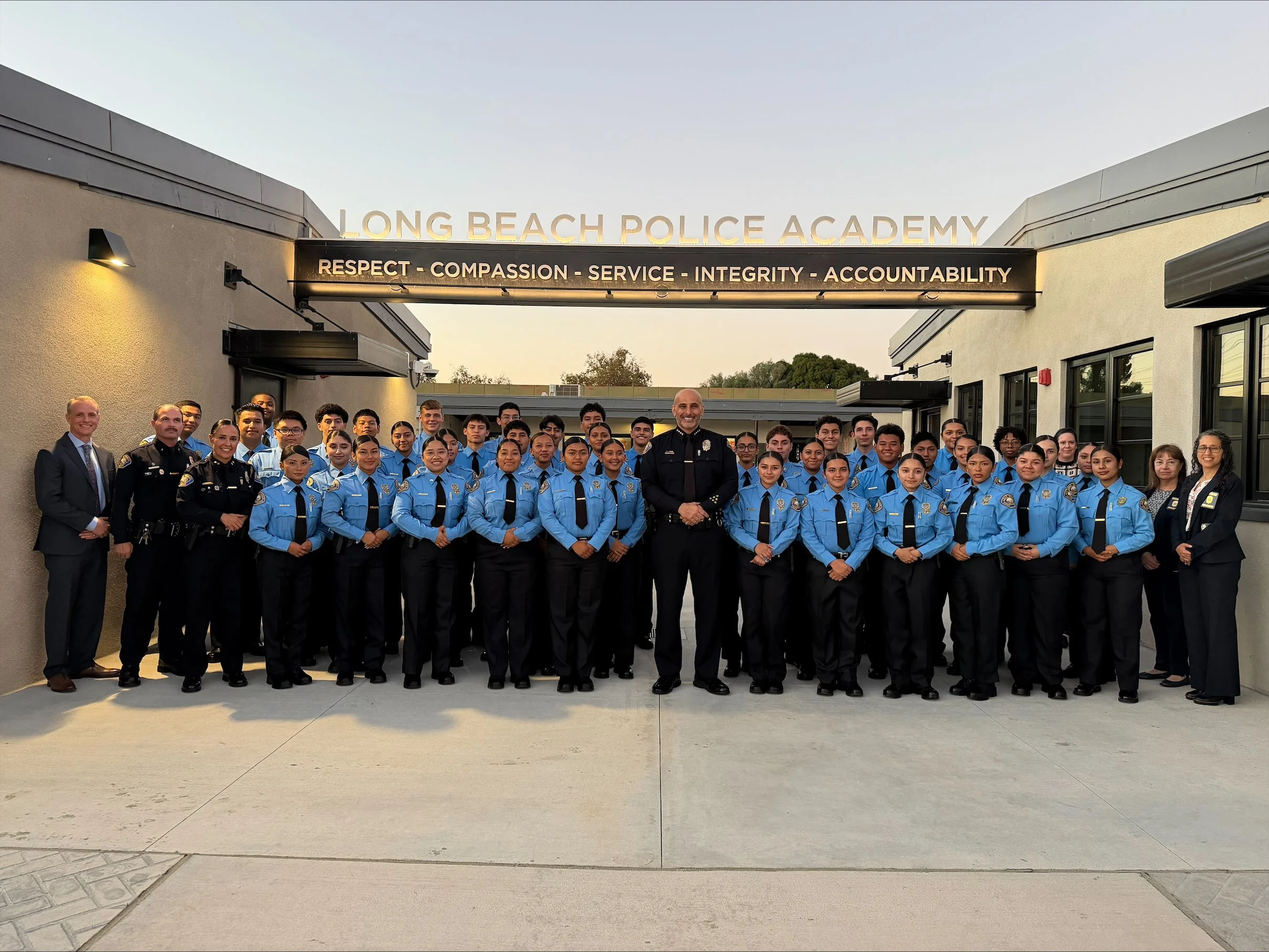 A diverse group of police officers, staff, and students from Long Beach Police Academy posing together outside the academy building, smiling for a group photo during daylight hours.