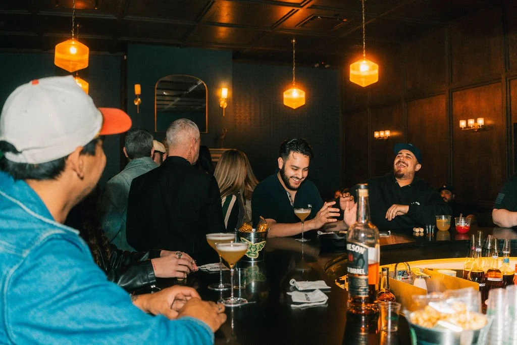 Many guests smiling and drinking at the bar of the Members lounge at Bike Shed Moto LA. With warm lighting.