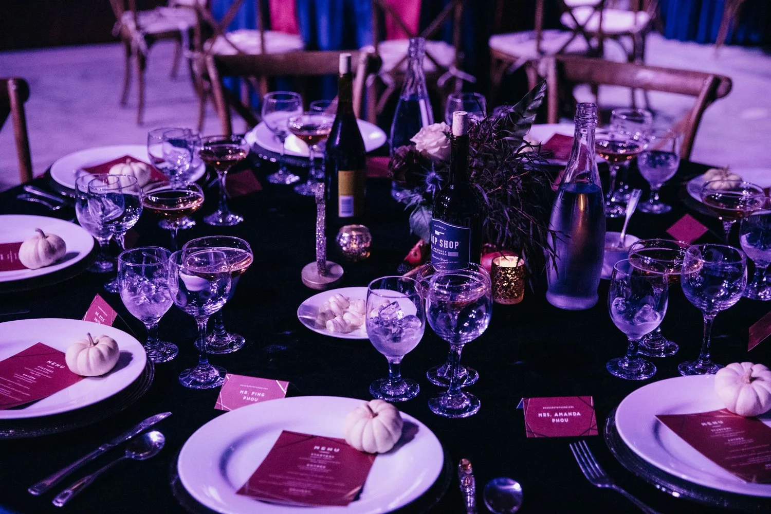 A round dining table set for a formal event with white plates, pink menus, small white pumpkins, multiple wine glasses, and water glasses. The table has a black tablecloth, and there are decorative items including bottles of wine, candles, and floral