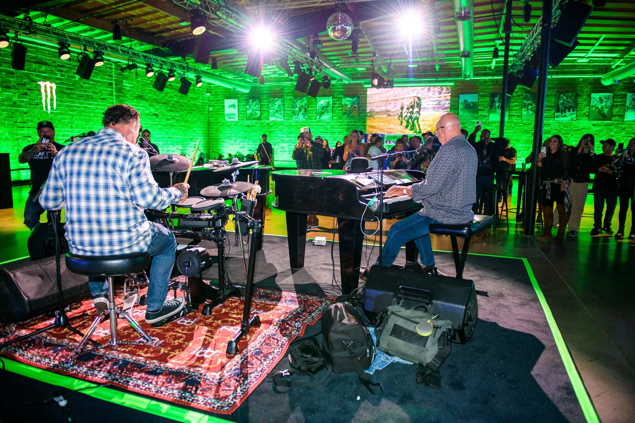 A trio band playing during an event to an audience at Bike Shed Moto LA, green lighting and rugs are on the stage