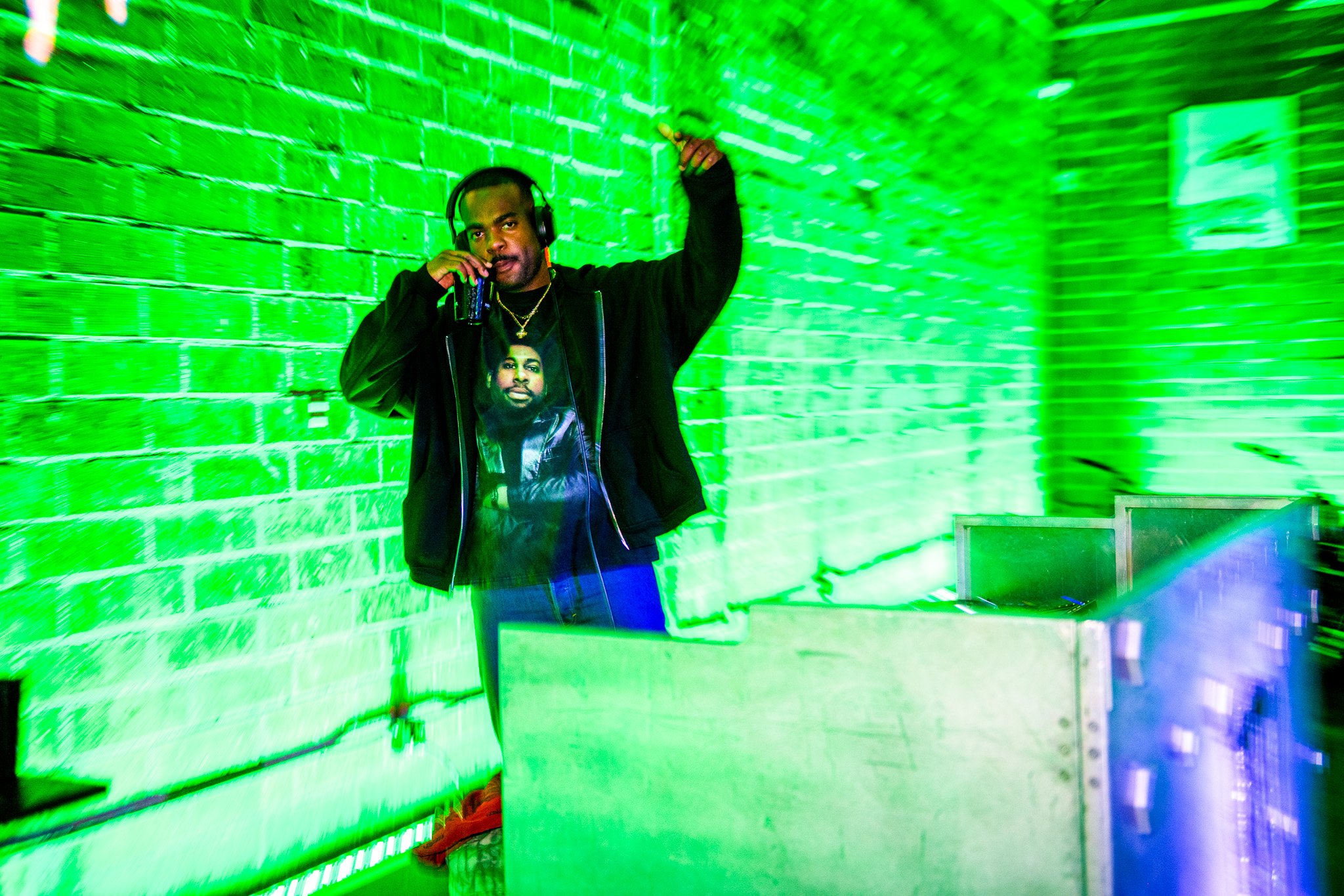 A moving blurring image of a DJ with his arm in the air and sipping on a drink during an event at Bike Shed Moto LA. Green lighting illuminated the scene and DJ Decks