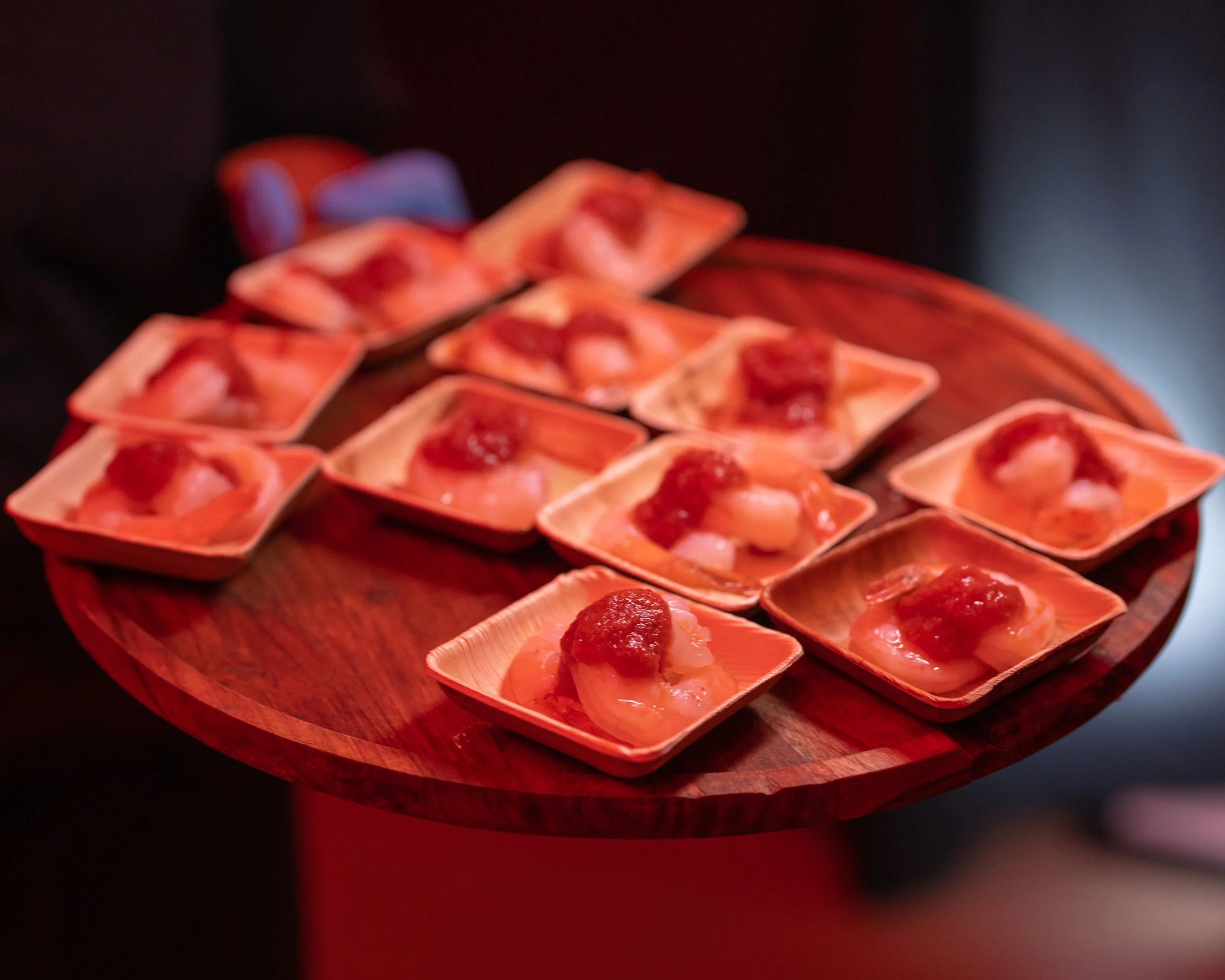 Small square paper plates with slices of fruit topped with red jam on a round wooden tray.