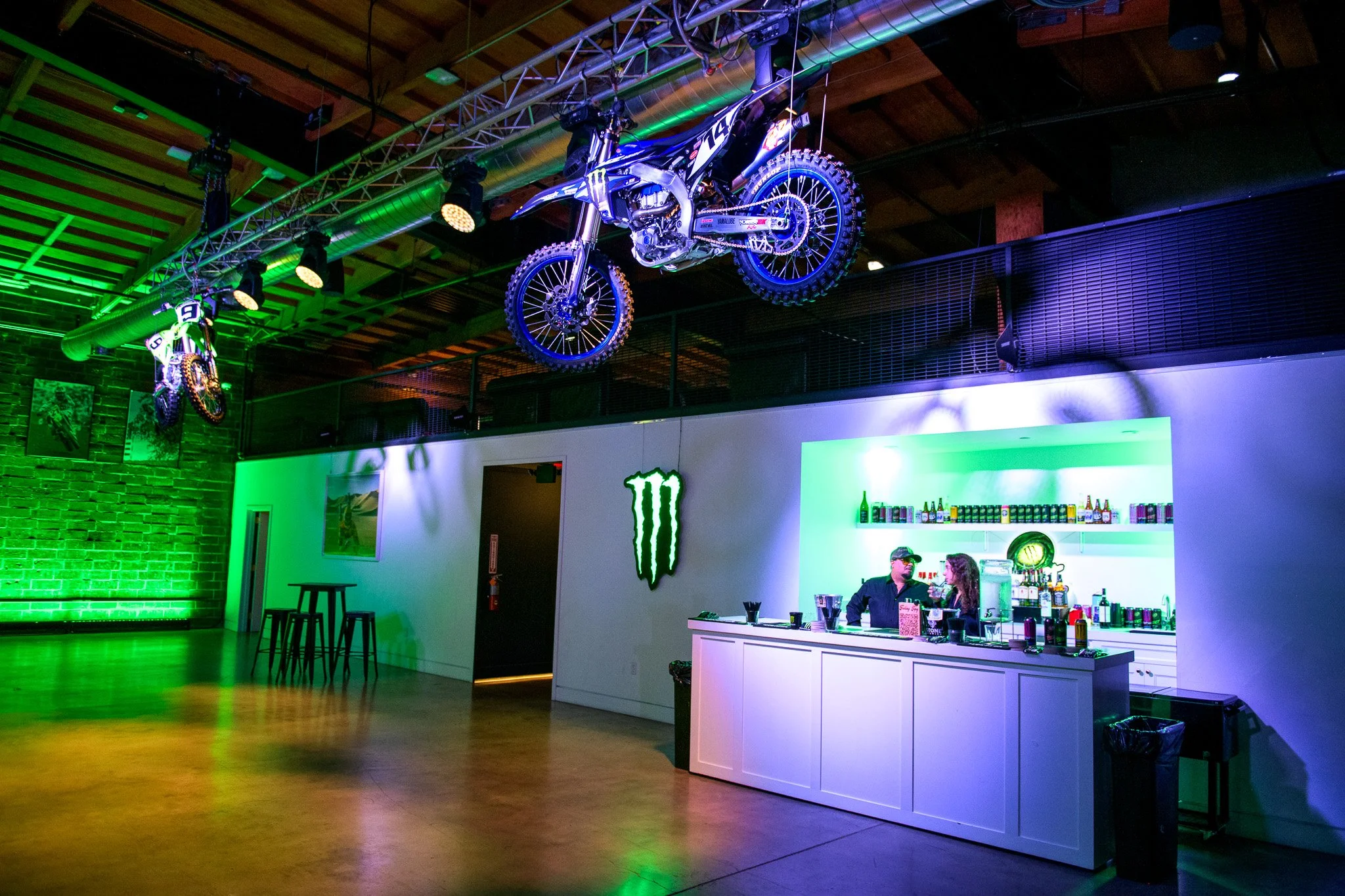 Motorcycle suspended in the air from metal railings at a Monster energy event with a bar and bartenders in the background, green lighting around
