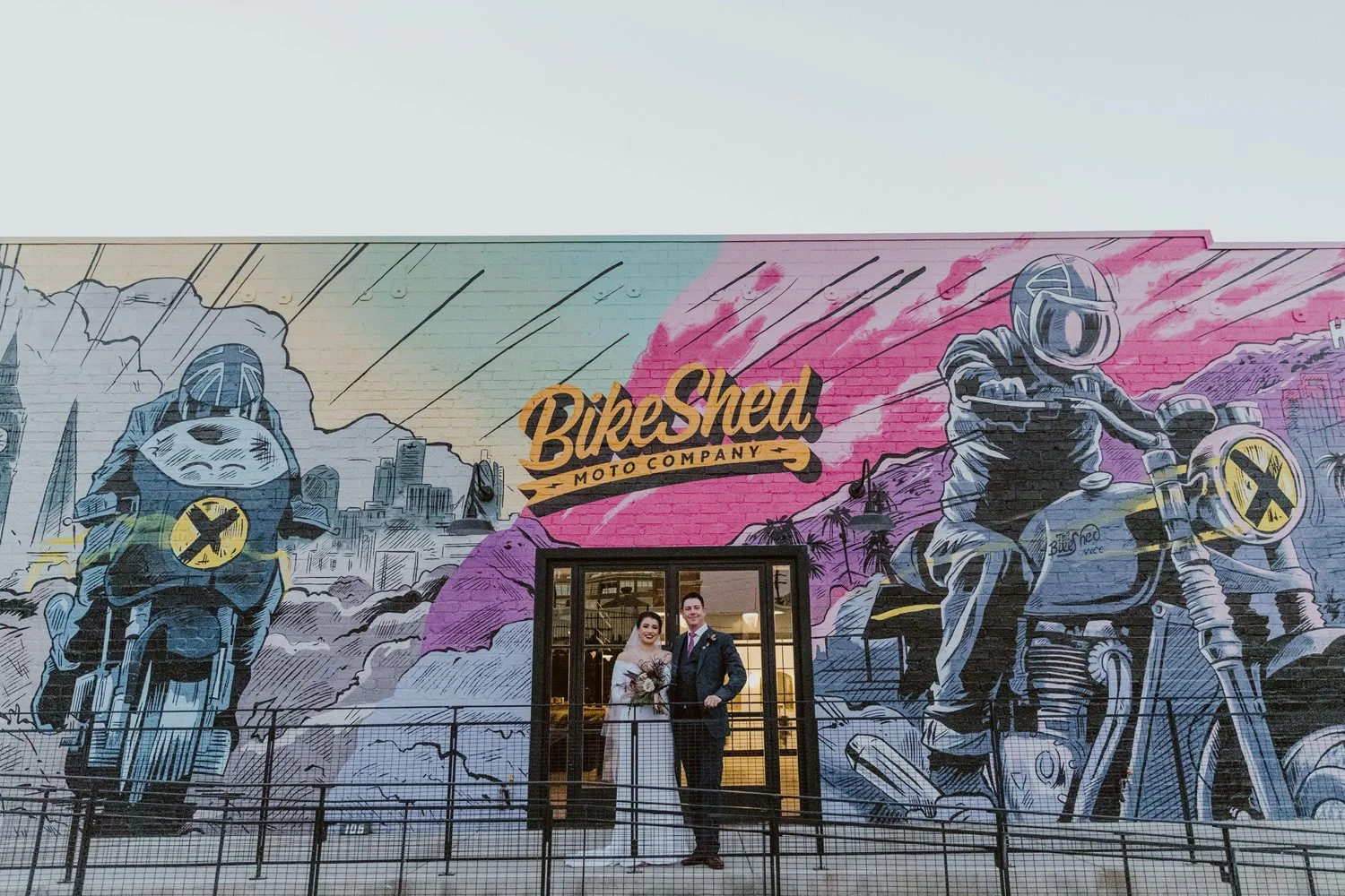 A mural featuring moto-themed artwork with two motorcyclists and the text 'Bike Shed Moto Company.' In front of the mural, a bride in a white wedding dress and a groom in a suit stand together, smiling, with the bride holding a bouquet.