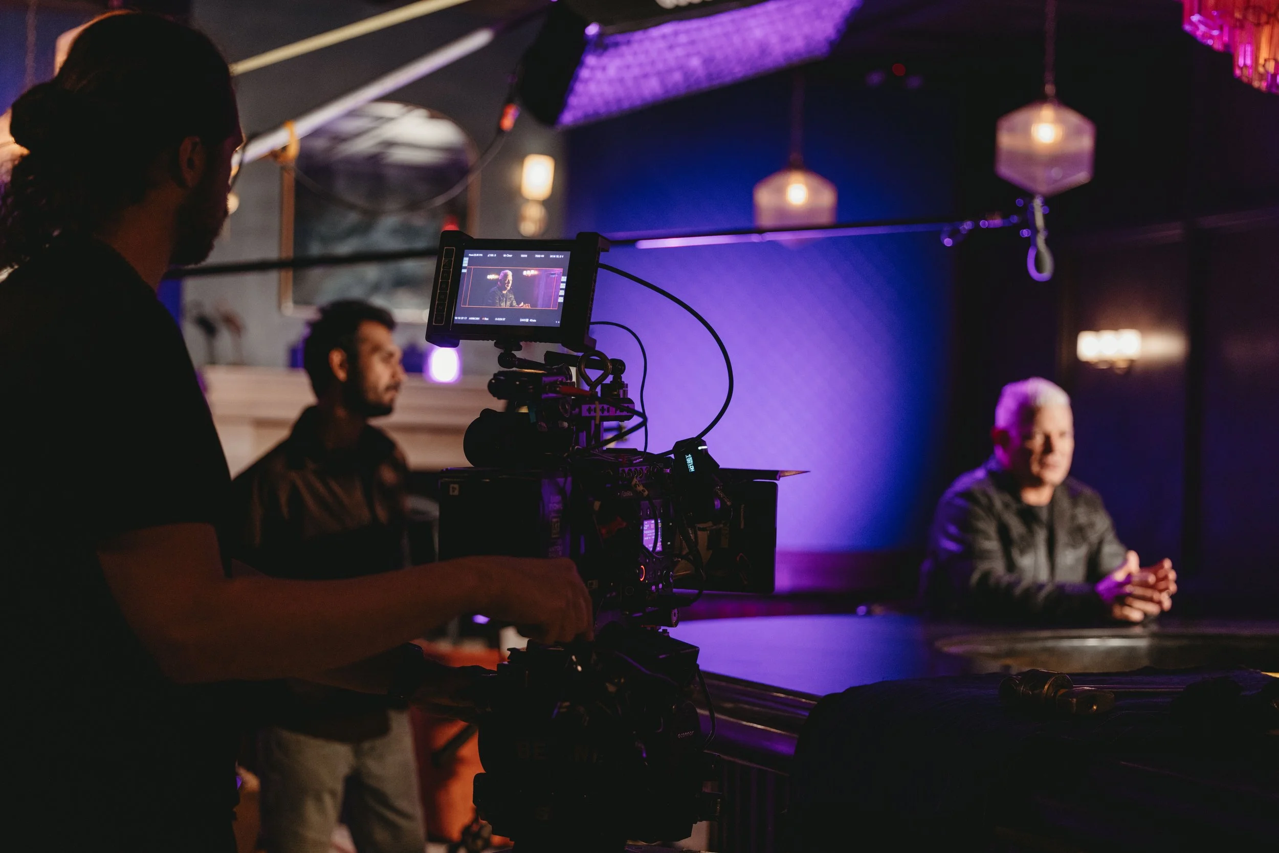 Man sitting a the bar being filmed for an event. Dim lighting and video camera are in shot