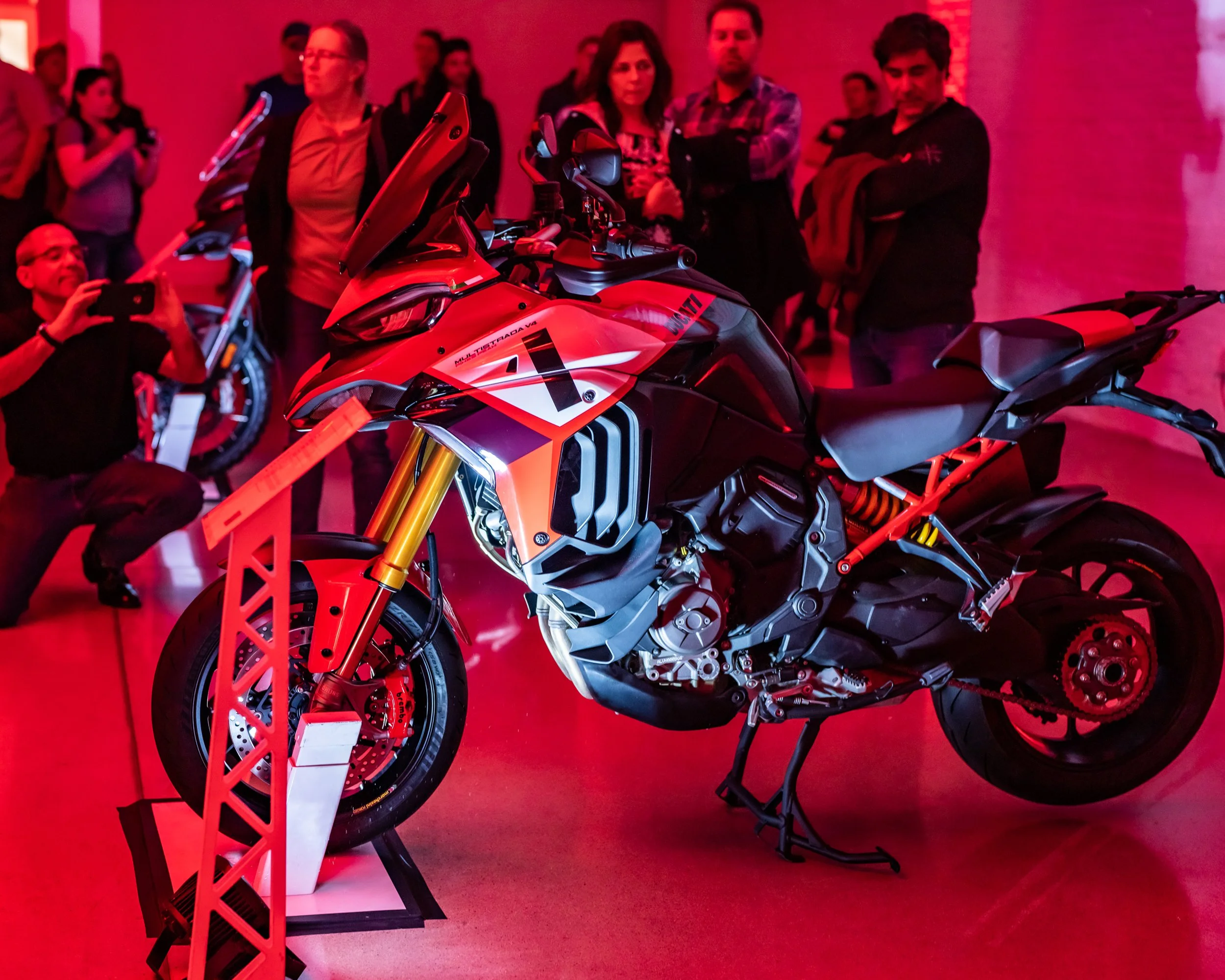 A red and black Ducati motorcycle on display at an event, with several people observing and taking photos in the background, illuminated by red lighting.