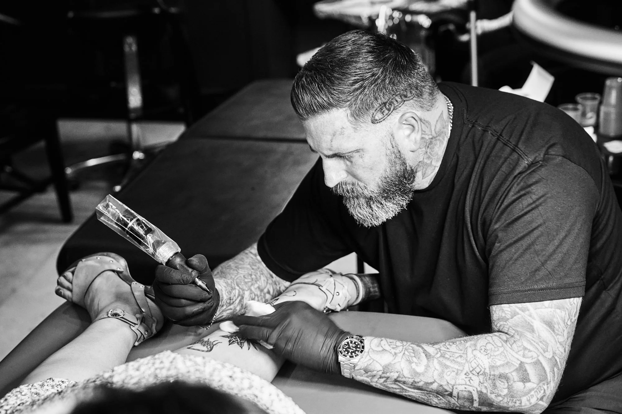 A tattoo artist with a beard and tattoos on his arms, wearing gloves, is working on a client's leg, using a tattoo machine in a studio setting.