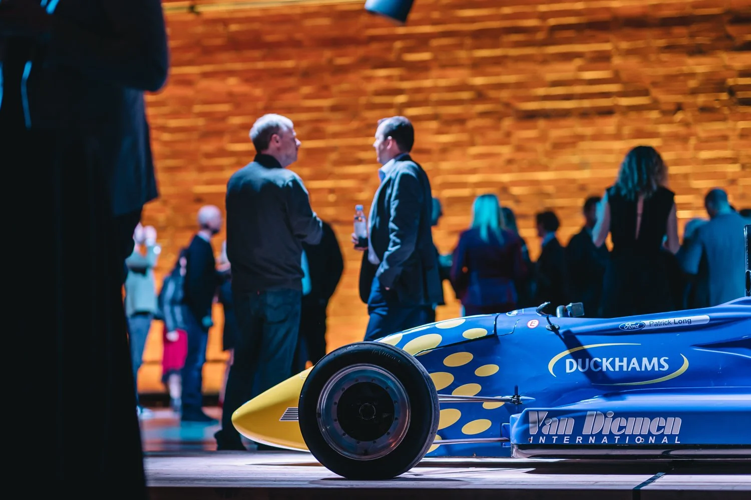 A photo of a blue racing car with yellow polka dots and the words "Duckhams" and "Van Diemen International" on it, displayed indoors on a stage or platform, with people in formal and casual attire conversing in the background.
