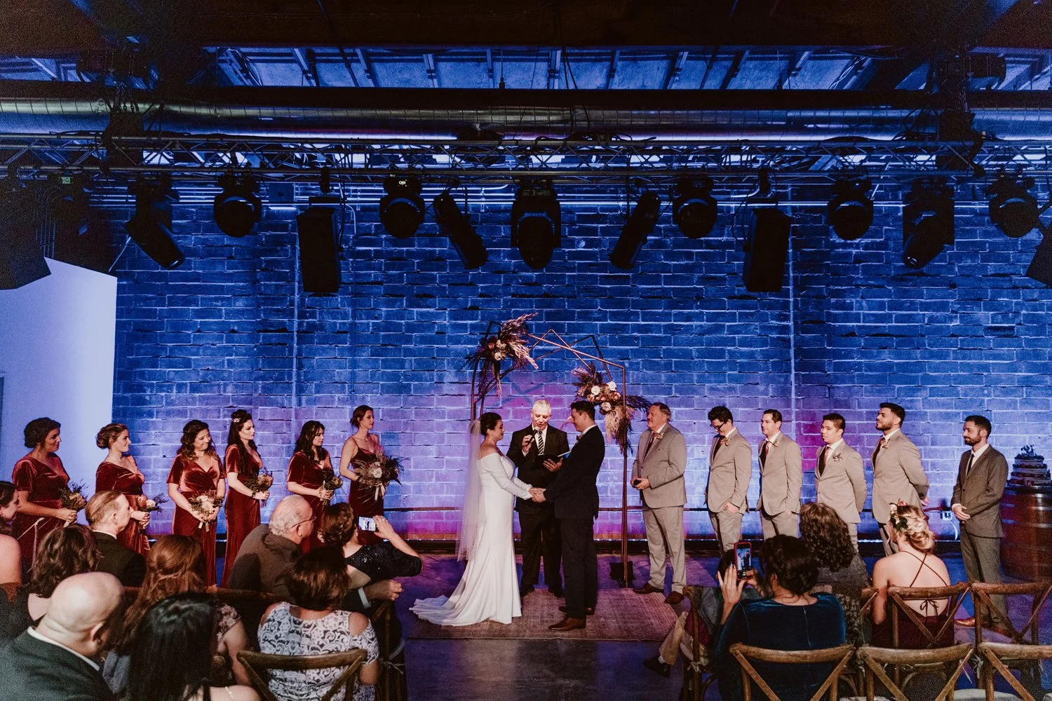 A wedding ceremony with a bride and groom exchanging vows on a stage, surrounded by their wedding party and guests, in a venue with exposed brick walls and stage lighting.