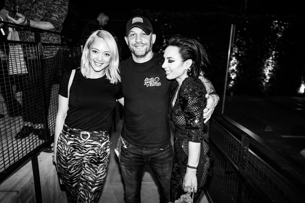 Three people smiling and posing for a photograph at an outdoor event at night, with a metal railing and trees in the background.